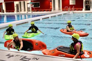 Five students sit in kayaks in an indoor swimming facility