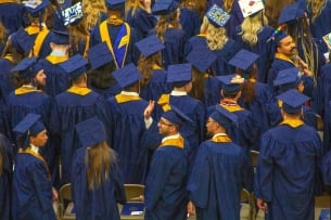 A group of students in blue and yellow graduation regalia face away from the camera. One student turns to the left to wave to someone out of frame.