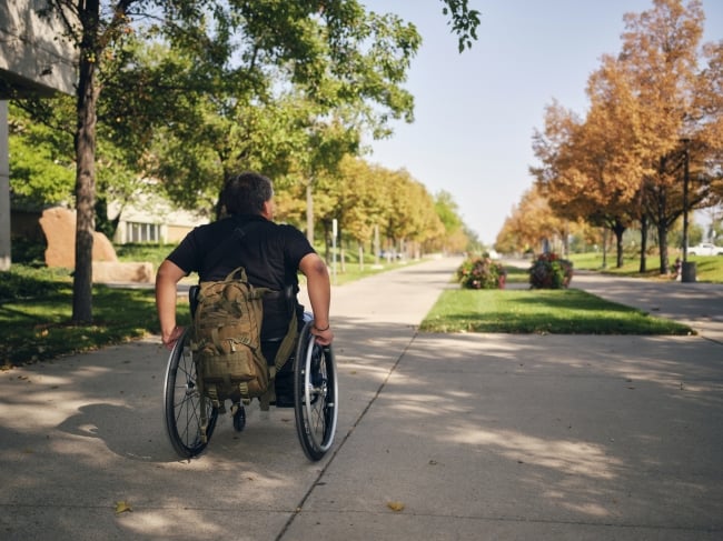 An adult disabled man, attending college, with a camo backpack hanging from his wheelchair.