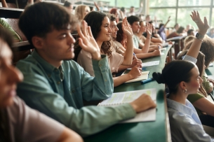Large group of university students raising their hands to ask the question on a class in amphitheater. Focus is on smiling woman.