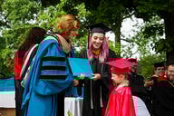 A mother and her young son, both dressed in graduation regalia, accept a diploma on stage from an administrator