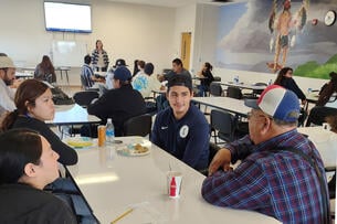 Students sit around a table talking with a Native American mural in the background.