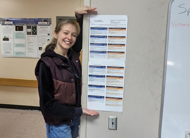 A young woman with blond hair holds up a white document with blue and orange boxes in a classroom.