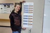 A young woman with blond hair holds up a white document with blue and orange boxes in a classroom. 